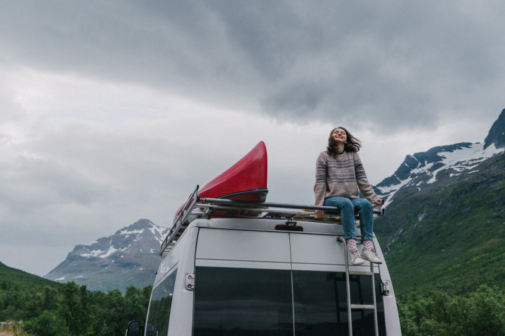 Young woman sitting on top of white van with a roof rack and red canoe